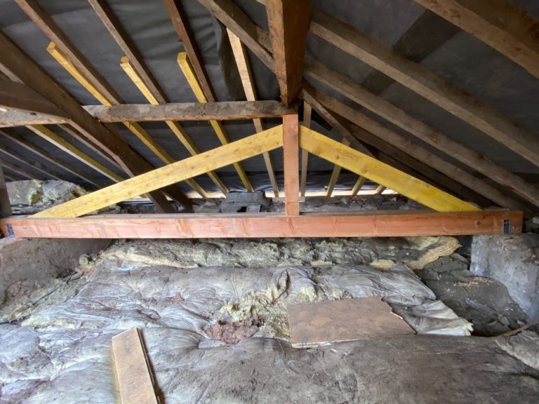 Vue d’un grenier montrant une charpente en bois rénovée avec des renforts en bois clair et des éléments isolants présents sur le sol.